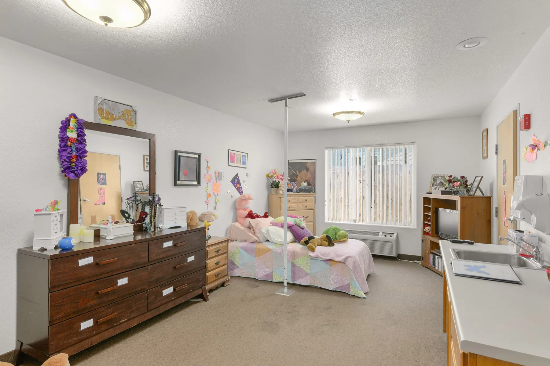 interior-10-1 (1) A cozy bedroom with a twin bed, colorful quilts, wooden furniture, and bright decor, featuring a window with blinds and a small kitchenette
